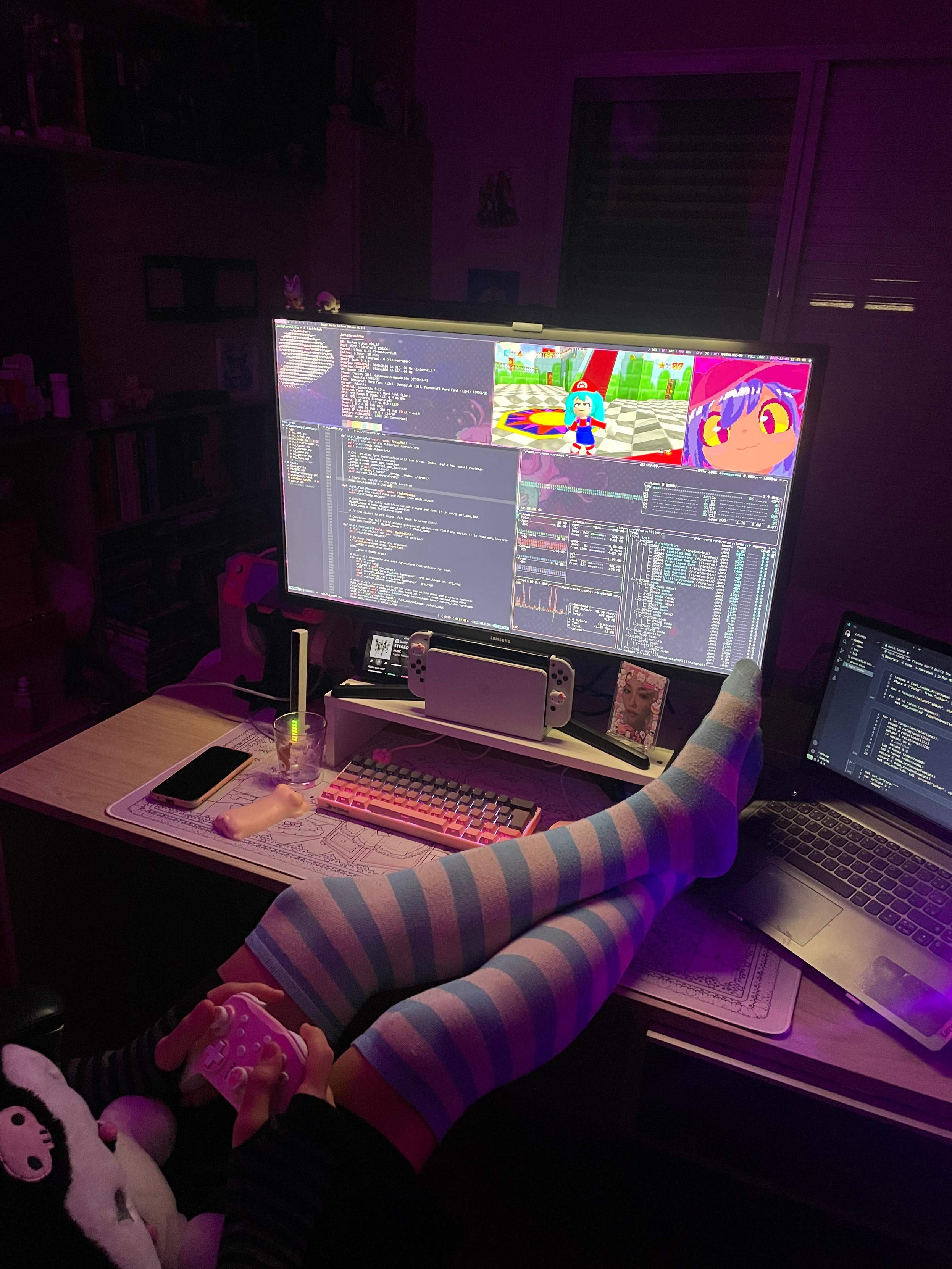 Person holding a Kuromi plush and a pink controller, wearing striped thigh high socks and resting their legs on a desk with a large monitor and a laptop running Gentoo Linux with bspwm. On screen there is code, btop, fastfetch, Niko from Oneshot and multiplayer SM64 with a Hatsune Miku skin.