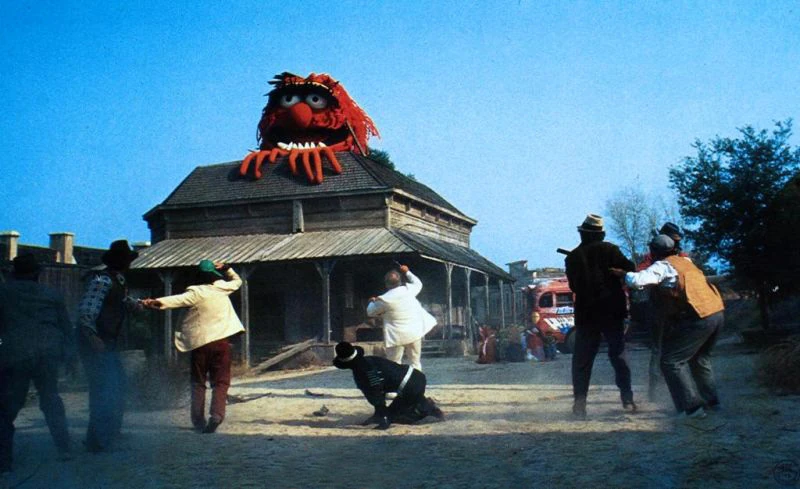 numerous people scramble at the site, one invoking what appears to be a shotgun, at the sight of a giant muppet head laying on the roof of a house